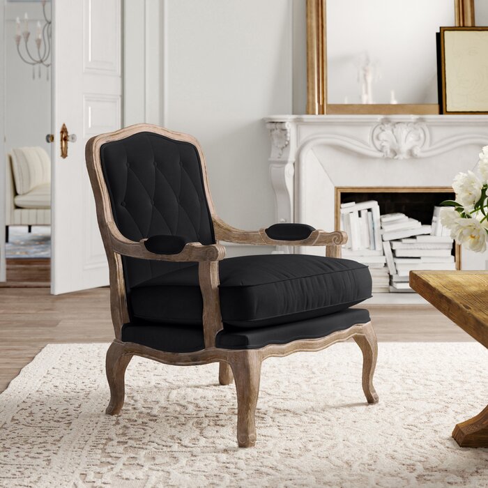 A black upholstered Sidqa Vintage Elegance Armchair with a distressed French wooden frame, placed in a room with a white rug and fireplace in the background.