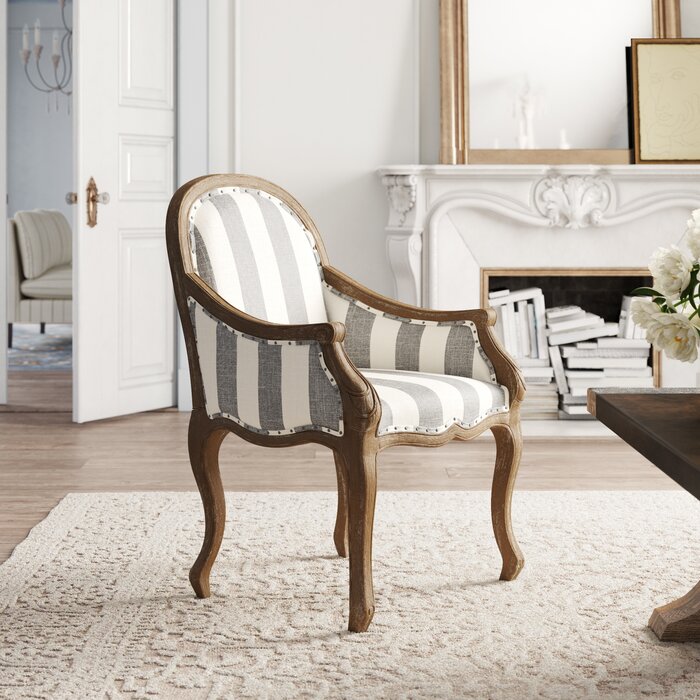 A gray and white striped armchair with a wooden frame, placed in a room with a white fireplace and a bookcase.