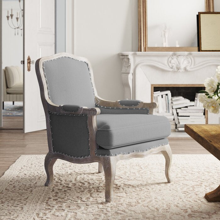 A gray upholstered armchair with a distressed wooden frame, nailhead trim, and a plush backrest and seat, positioned in a room with a classic decor.