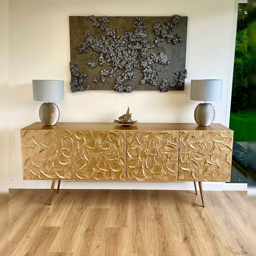 An elegant sideboard with hand-carved petals in a soft gold finish, slim golden legs, and a decorative painting above it in a contemporary design style.