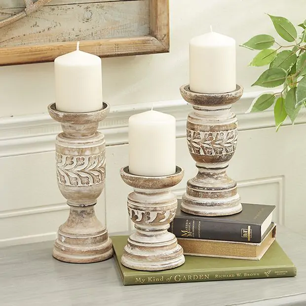 A set of three white wash wooden candle stands with an ornate foliage design, holding white candles, placed on a surface with a green book and a plant in the background.
