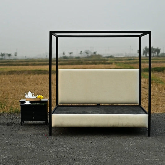 A black metal canopy bed with cream upholstered headboard and footboard, shown outdoors on a paved surface beside a small black side table in an open field under a hazy sky.