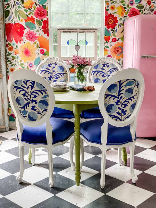 A vibrant dining set featuring a green table with a floral pattern on the chairs and a blue cushion seat, set against a colorful wall with floral wallpaper.