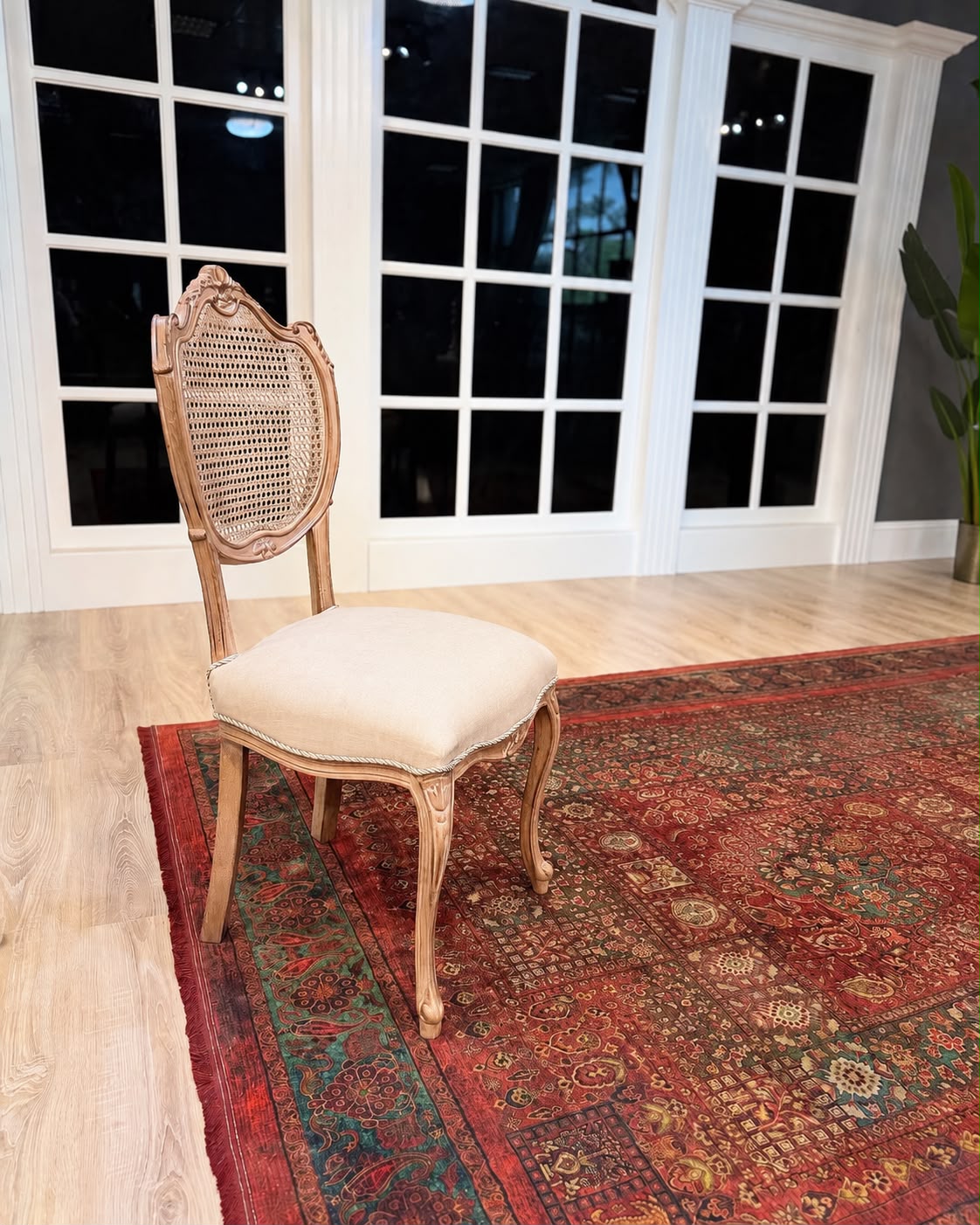 A classic French-style dining chair with a carved light wood frame, woven cane backrest, and cream upholstered seat cushion, positioned on a red patterned area rug in front of white French doors.
