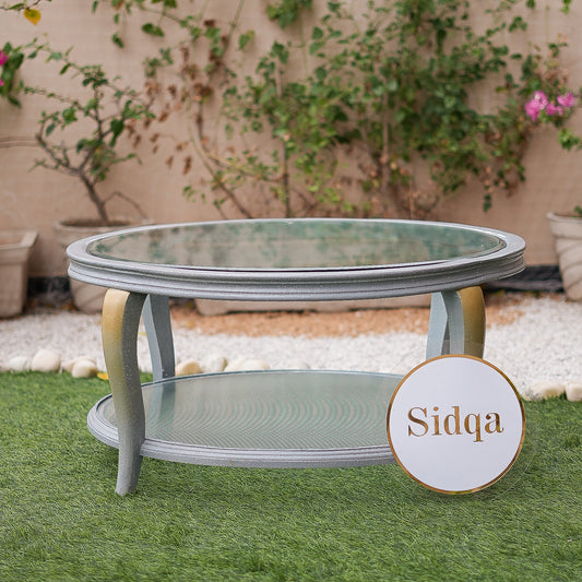 Round glass table with metal frame on grass, 'Sidqa' brand visible