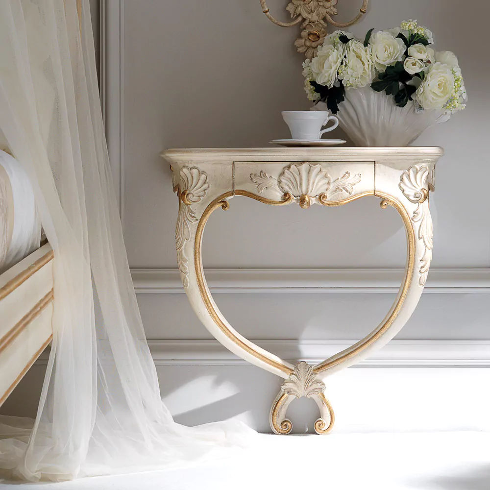 An ivory wall mounted console table with gold carvings, displayed in a room setting with a cup and saucer on top, alongside a floral arrangement.