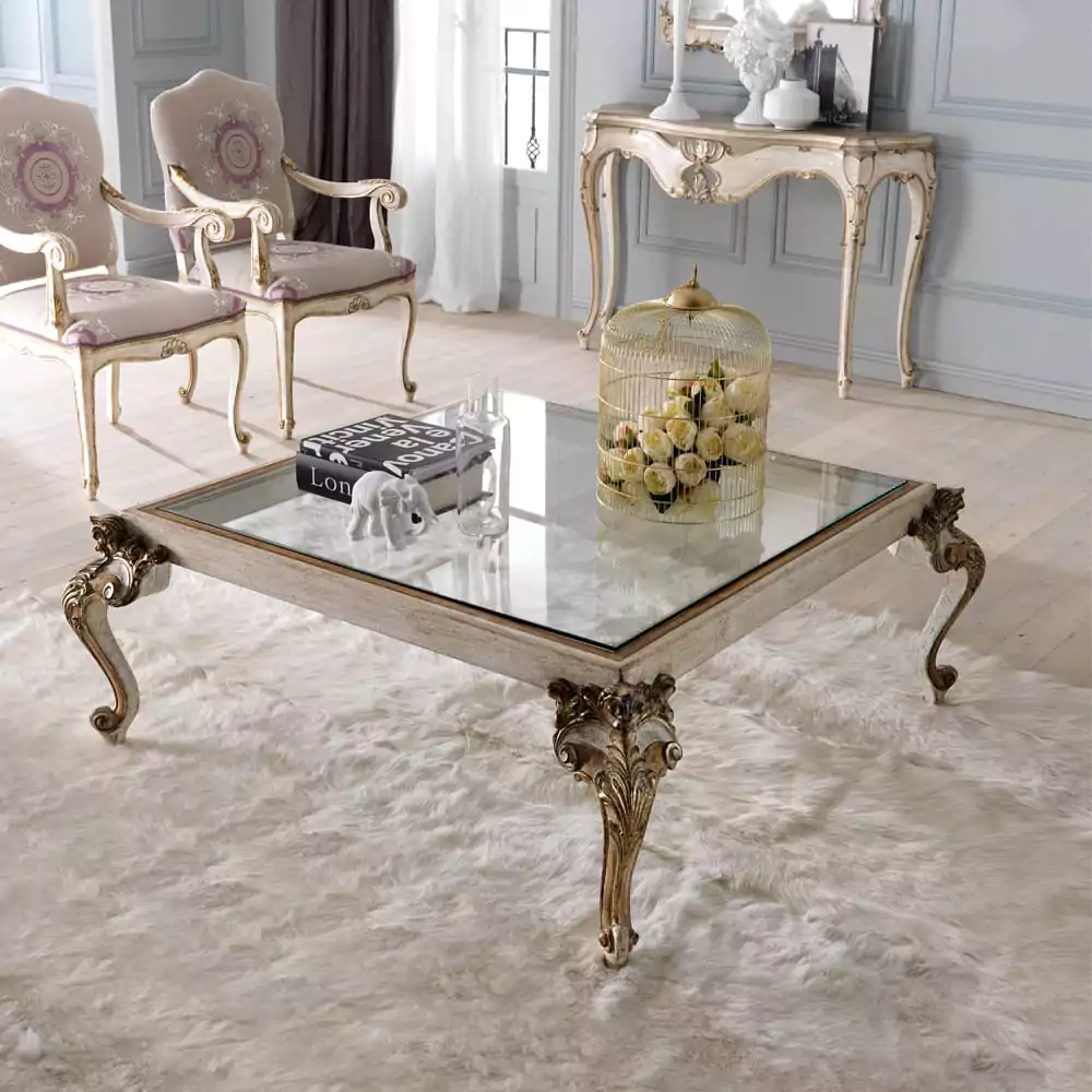 A luxurious square glass-top coffee table with gold brown carvings and thick feet, placed on a white furry rug in a stylishly furnished living room.