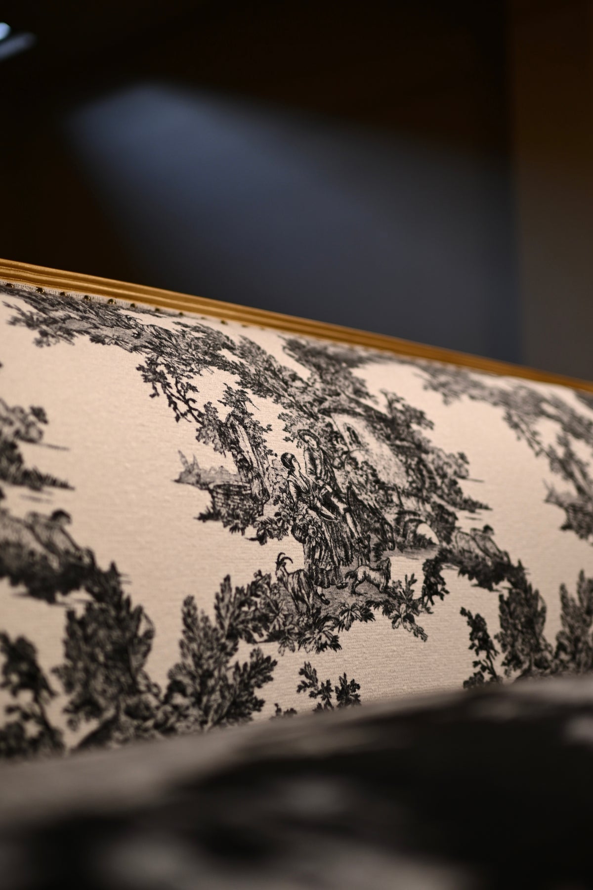 Close-up of a sofa cushion upholstered in black toile pattern fabric on a cream background, with a gold-toned frame edge visible at the top.