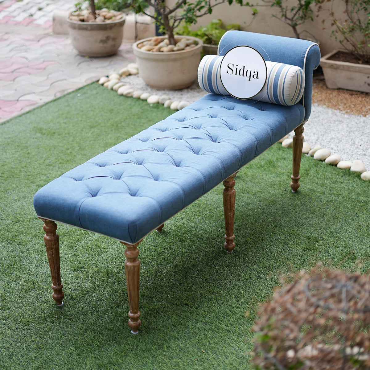 Blue bench with tufted backrest and wooden legs on a grassy area with potted plants.