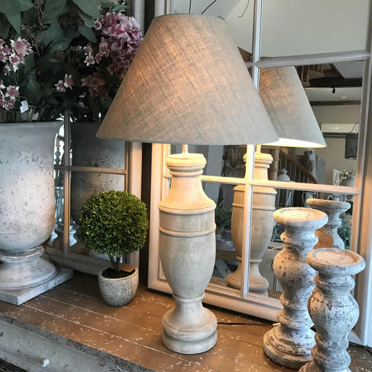A beige Surahi Elegance French Finish Table Lamp with a traditional shape, displayed in a room setting with decorative items and a mirror in the background.