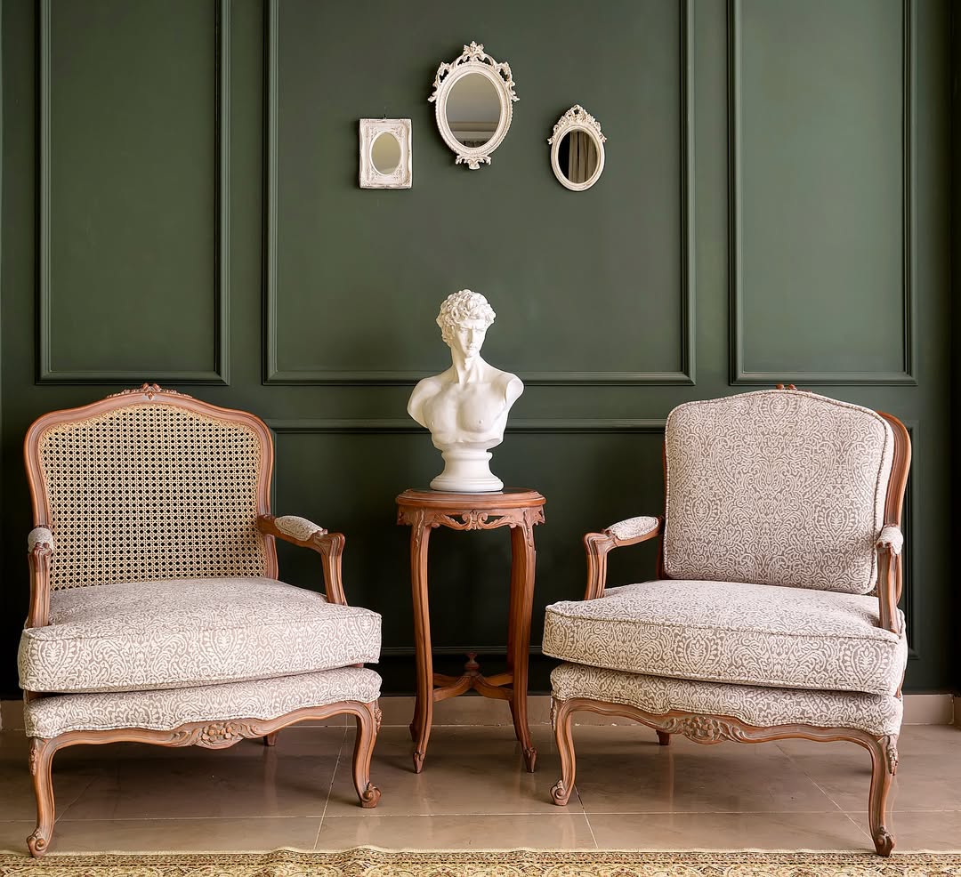 A luxurious beige upholstered 3-seater sofa with cane detailing and teak wood legs, accompanied by two patterned pillows, positioned against a dark green wall with decorative mirrors above.
