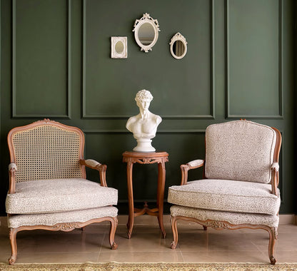 A luxurious beige upholstered 3-seater sofa with cane detailing and teak wood legs, accompanied by two patterned pillows, positioned against a dark green wall with decorative mirrors above.