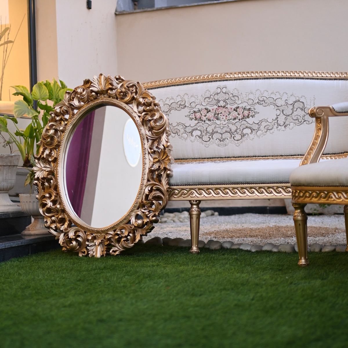 Decorative gold mirror and chair in front of a white sofa with floral patterns on a grassy area.