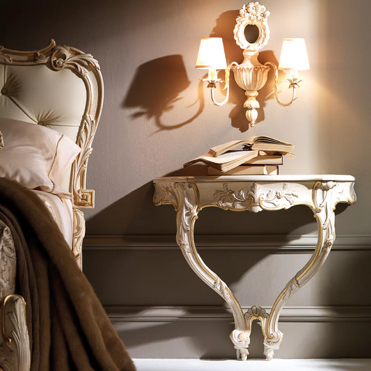 An ivory French wall-mounted console with intricate carvings and gold accents, displayed in a room setting with a lamp and mirror on top.