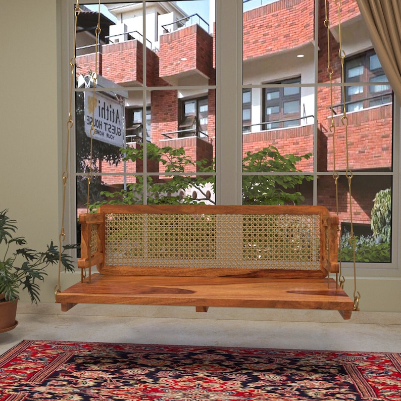 A wooden swing with intricate cane backrest, suspended by golden steel hangers in an indoor setting with a large window in the background.