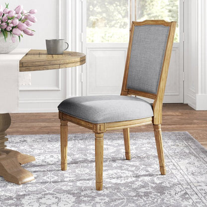 A contemporary rectangular chair with light grey fabric upholstery and wooden legs, placed in a room with a round wooden table and a floral arrangement.