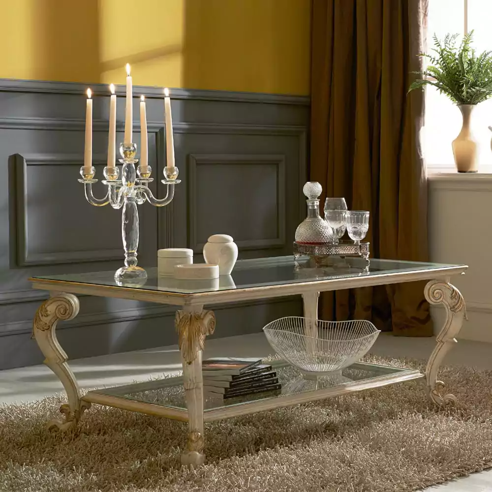 An ivory and gold glass double-decker coffee table with ornate legs, placed on a carpeted floor in a room with a yellow wall and a black chair. The table has decorative items on it, including a crystal candelabra and glasses.