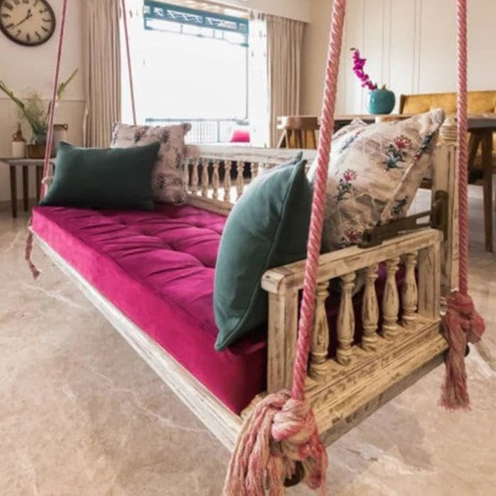 Wooden swing with pink cushion and green pillows in a room with a clock and table in the background.