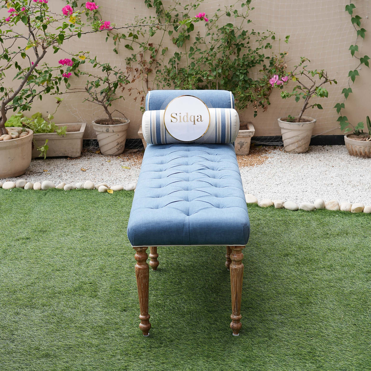 Blue tufted bench with wooden legs on a grassy area with plants in the background