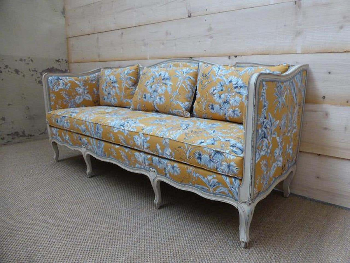 A yellow floral upholstered sofa with a white distressed French wooden frame, positioned against a wooden backdrop.