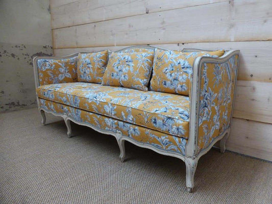 A yellow floral upholstered sofa with a white distressed French wooden frame, positioned against a wooden backdrop.