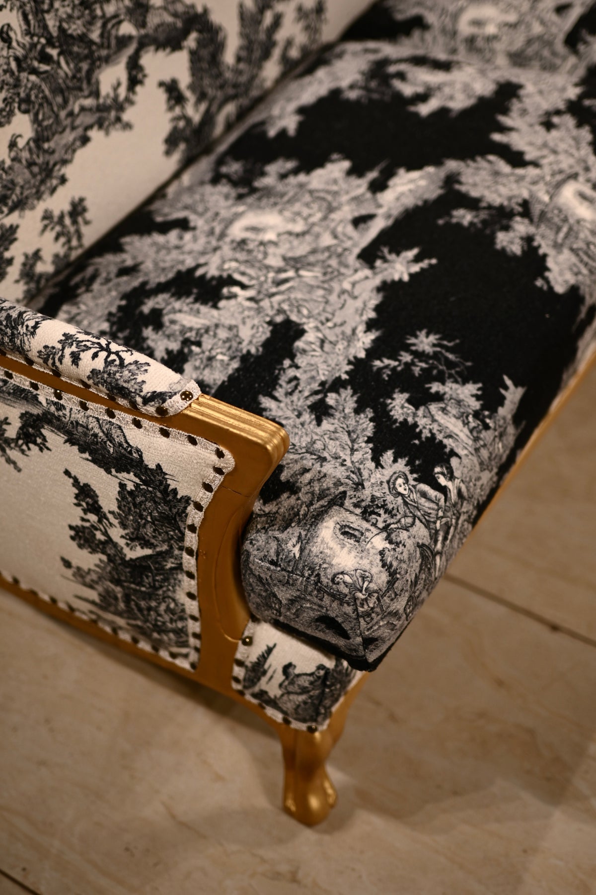 Close-up of an upholstered armchair with black and white toile pattern fabric and a gold wooden frame, shown on a light stone floor.