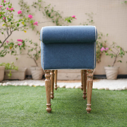 Blue upholstered stool with wooden legs on a grassy area with plants in the background