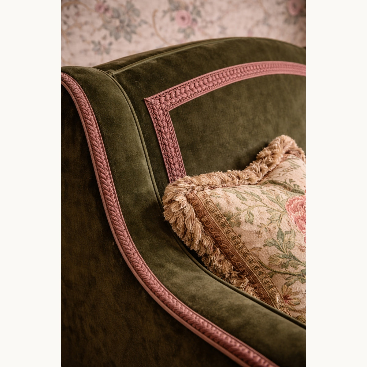 Green velvet armchair with pink trim and a patterned pillow against a floral wallpaper background