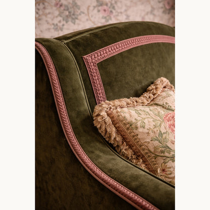 Green velvet armchair with pink trim and a patterned pillow against a floral wallpaper background