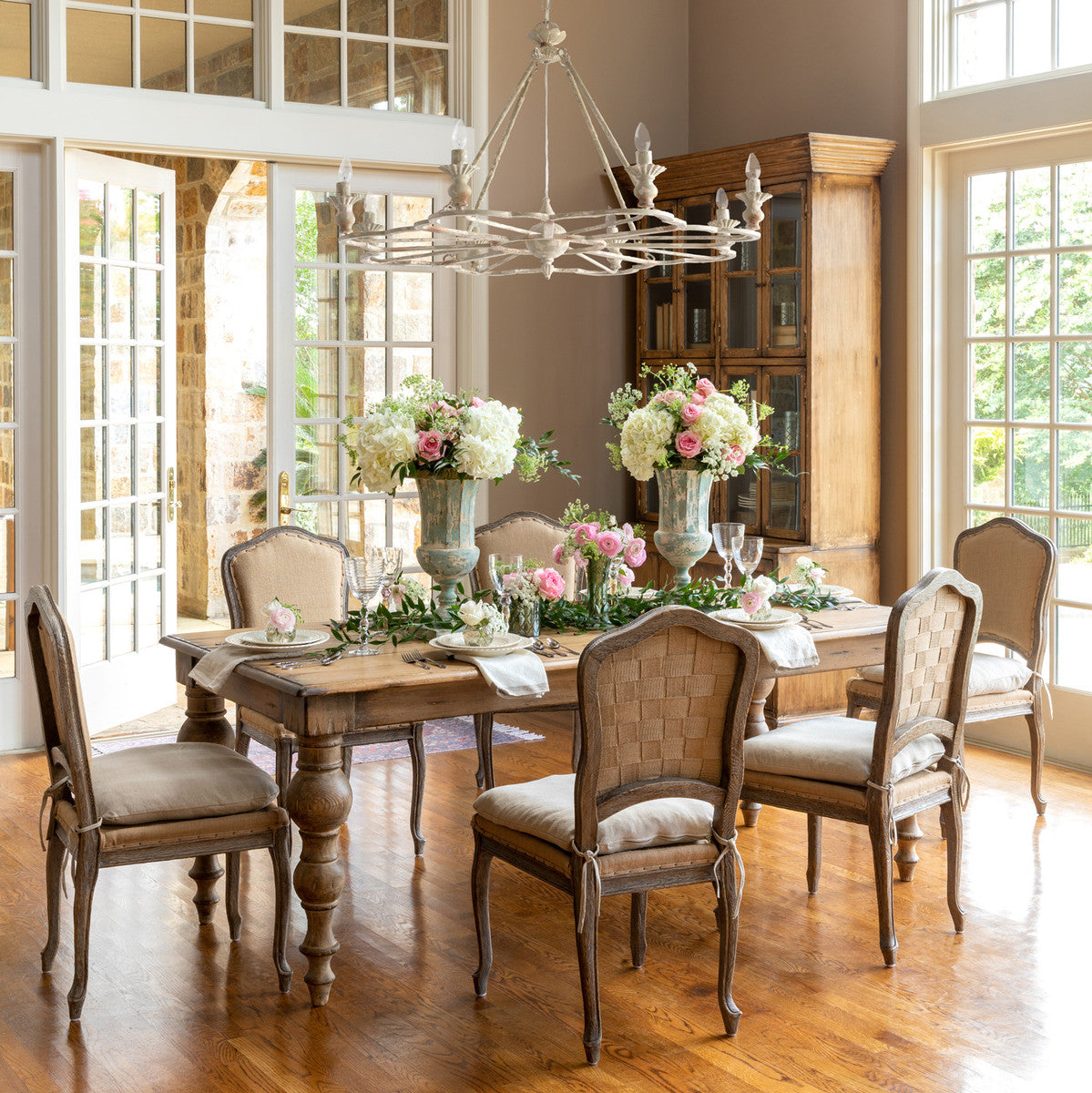 A set of elegant dining chairs with beige fabric upholstery and ivory cushions, around a wooden dining table, in a room with a chandelier and large windows.
