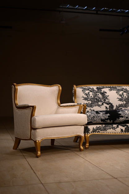 An elegant armchair with cream upholstery and gold-toned wooden trim, positioned next to a matching sofa with black and white toile pattern in a dimly lit showroom.
