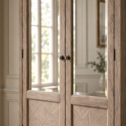 A rustic double-door cabinet or wardrobe with light weathered wood grain, geometric patterned lower panels, and dark metal knobs, shown in a bright interior room with a window and wall molding visible.