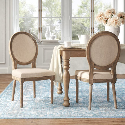 A beige fabric upholstered oval chair with a natural distressed wood frame, positioned in a room with a wooden dining table and large windows.