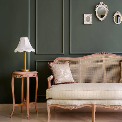 A luxurious beige upholstered 3-seater sofa with cane detailing and teak wood legs, accompanied by two patterned pillows, positioned against a dark green wall with decorative mirrors above.