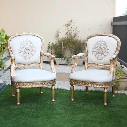 Pair of ornate chairs with gold frames and white cushions on a grassy area.
