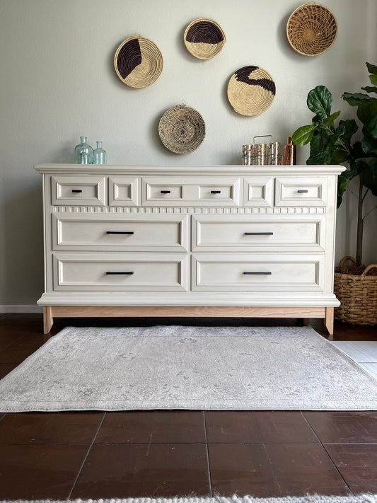 A white buffet cabinet with gold handles, placed against a wall with decorative plates, a plant to the side, on a grey area rug.