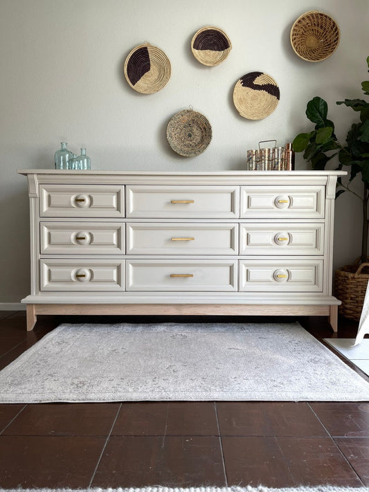 A white buffet cabinet with gold handles, placed against a wall with decorative items on top and a plant to the side, on a gray rug.