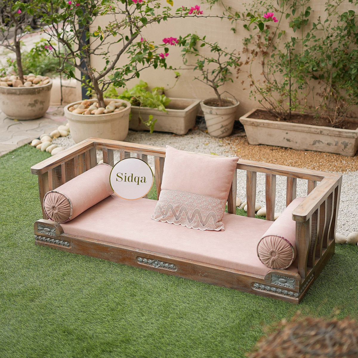 Wooden outdoor bench with pink cushions and 'Sidqa' branding in a garden setting.