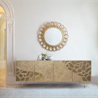 An elegant sideboard buffet table with a carved, textured surface in gold finish, balanced on sleek brass legs. A round mirror with a decorative gold frame hangs above the table, enhancing the luxurious feel of the scene.