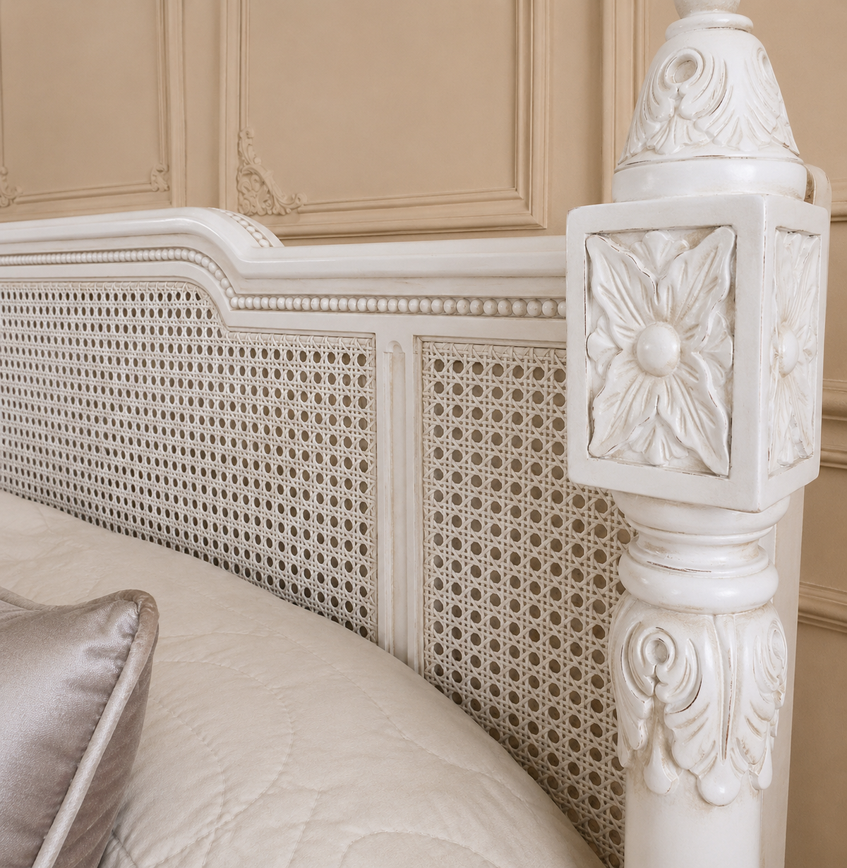 Close-up of an ornate white carved wooden bed frame headboard with woven cane panels, dressed with cream bedding and a satin pillow in a classic paneled room.