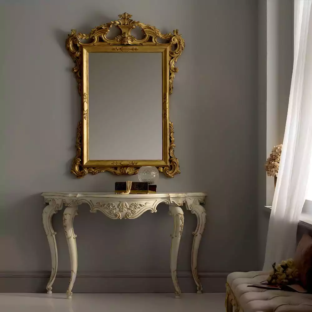 An ivory console table with gold accents on the carvings, accompanied by an antique gold mirror with intricate detailing, placed in a room with a gray wall and a hint of a floral curtain on the right.