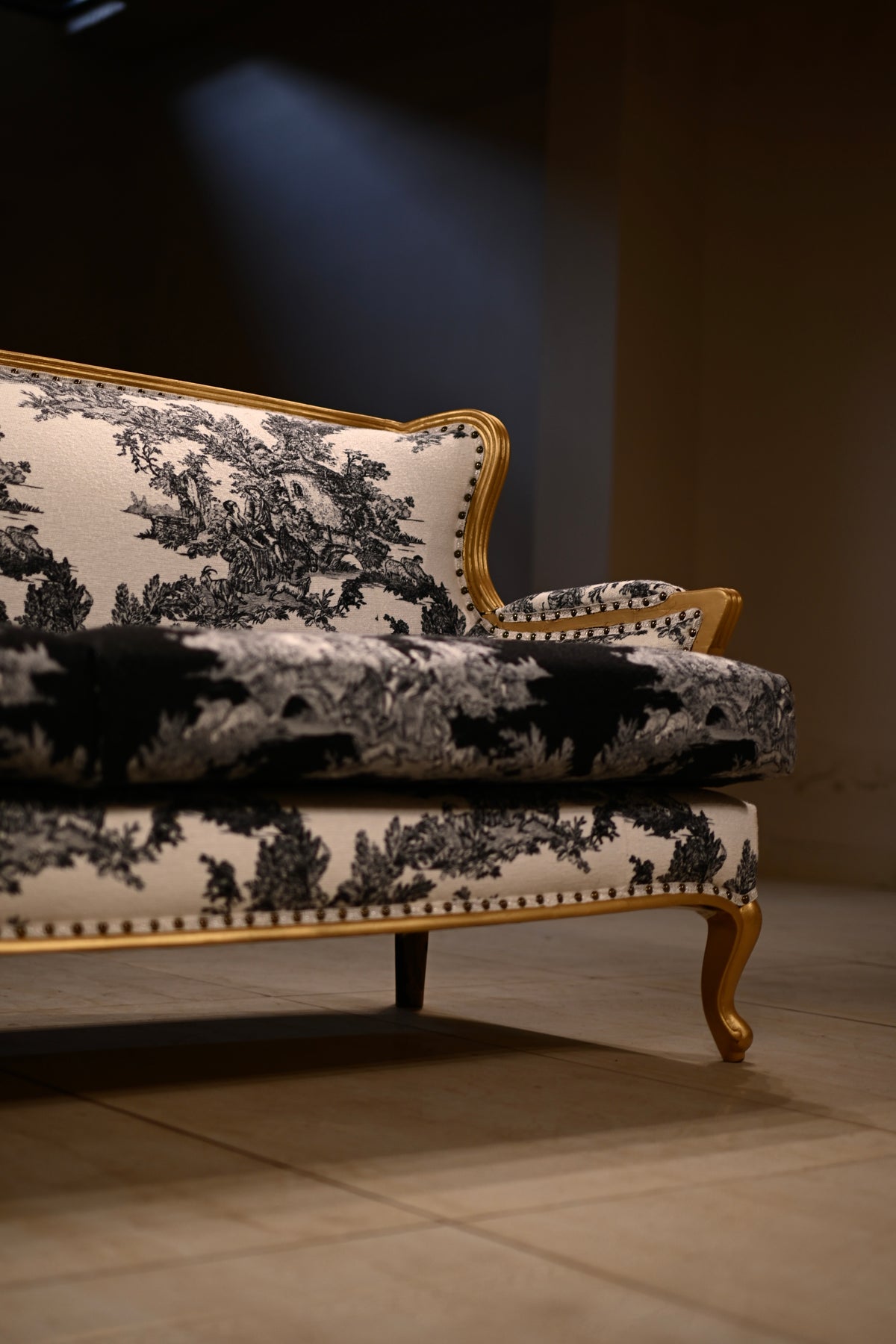 A close-up of a classic French provincial sofa upholstered in a black and white toile pattern with a gold wooden frame and nailhead trim, set against a dark background.