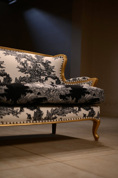 A close-up of a classic French provincial sofa upholstered in a black and white toile pattern with a gold wooden frame and nailhead trim, set against a dark background.