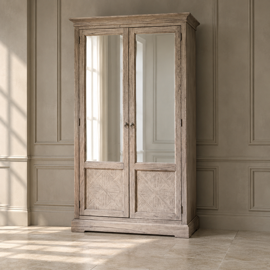 A tall rustic wooden armoire cabinet with two mirrored doors and carved lower panels, shown in a styled room with paneled walls and tiled floor.