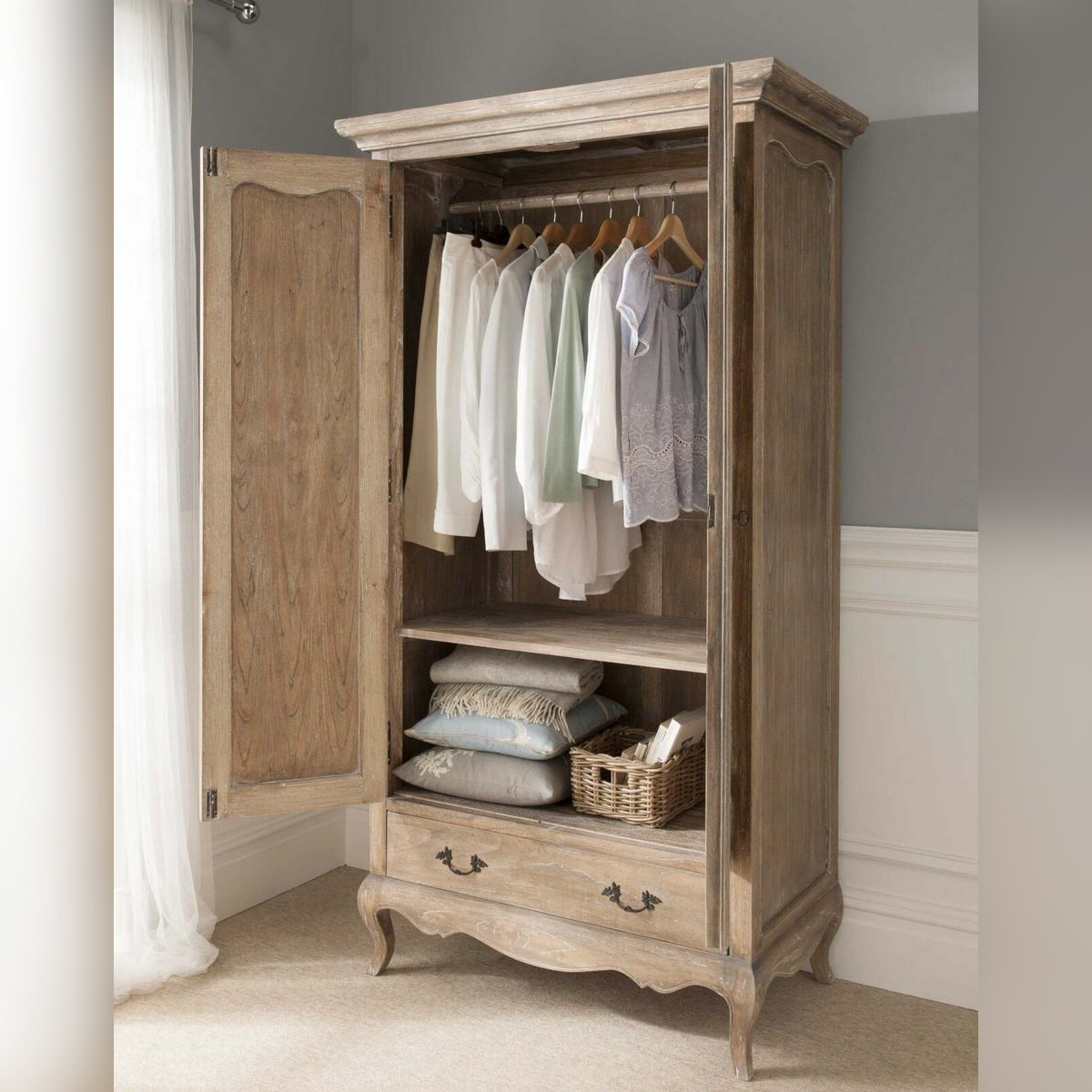 Wooden wardrobe with clothes hanging inside against a neutral wall.