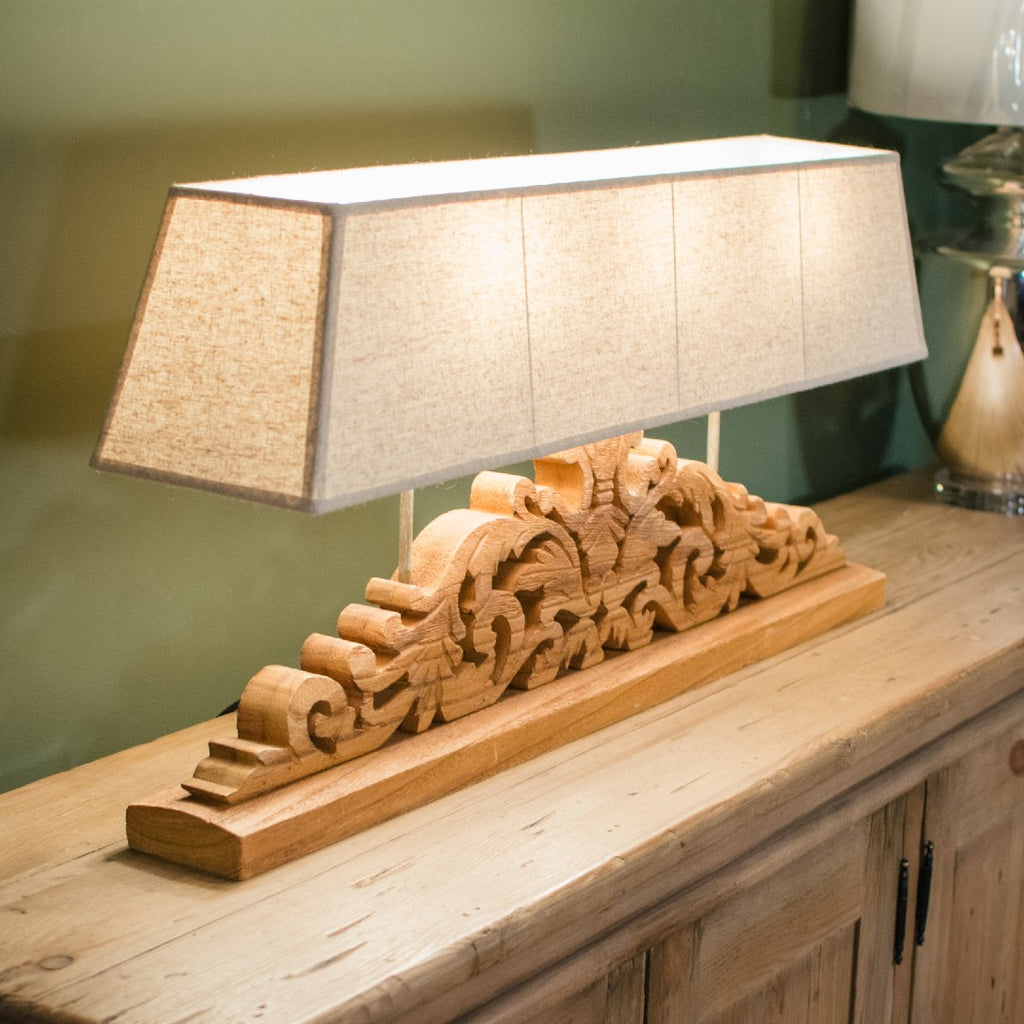 An intricately carved wooden table lamp with a wide rectangular shade, placed on a wooden surface.