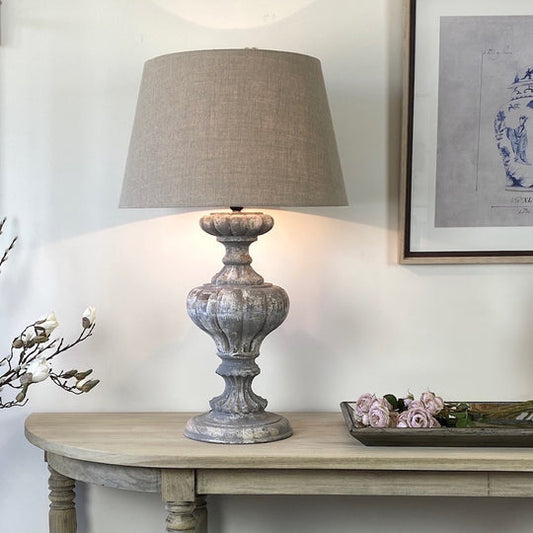 An elegant single table lamp with a French distressed grey finish, placed on a wooden table in a room with a neutral decor.
