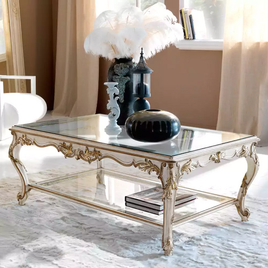 A luxurious double-decker coffee table with a white French finish and antique gold carvings, featuring glass tops and placed in a well-decorated living room setting.
