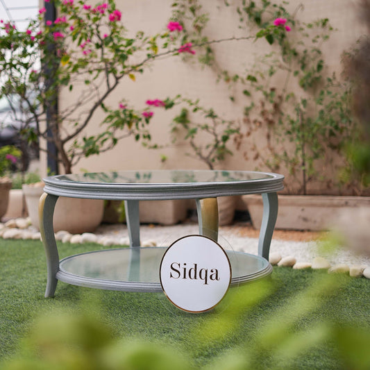 Outdoor setting with a glass coffee table and 'Sidqa' logo in the foreground.