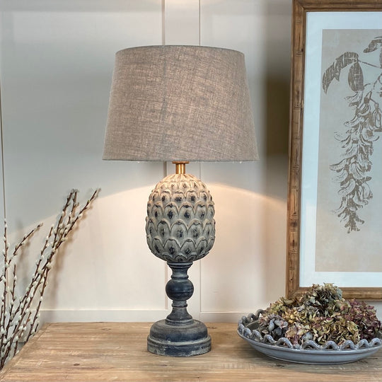 A decorative table lamp with a carved base resembling an pineapple, finished in a moody patina, and a natural shade. The lamp is placed on a wooden surface with a framed picture and a decorative bowl in the background.